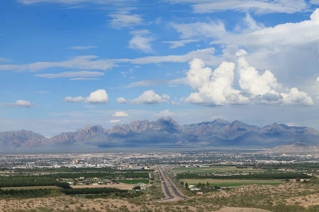 Aerial view of a road leading to a city surrounded by green fields, with a backdrop of mountains and a partly cloudy sky.