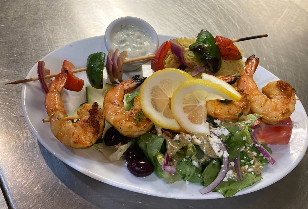 Grilled shrimp skewers with lemon slices, vegetables, and a side salad with feta cheese and olives, served on a white plate at Kallos Mediterranean Cuisine.