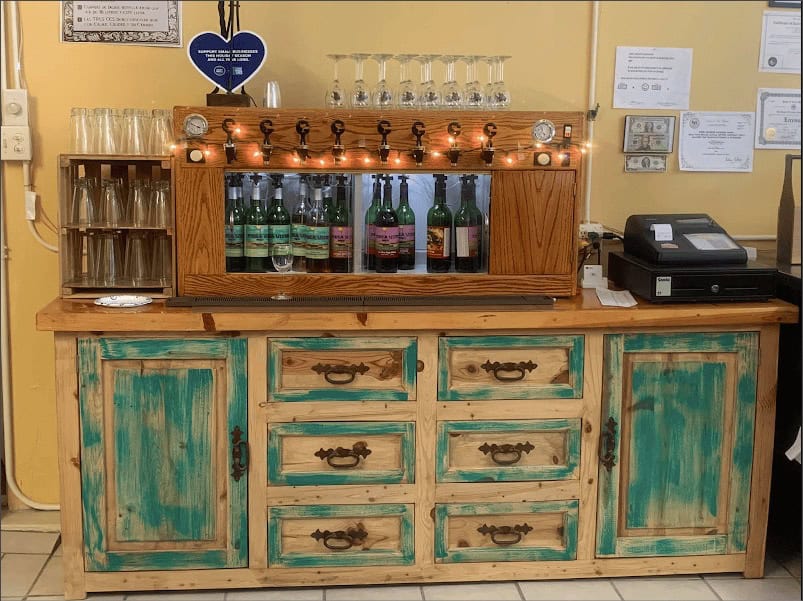 A rustic wooden counter in the Winery Tasting Room with distressed teal cabinets, displaying wine bottles and glasses, lit by string lights above.