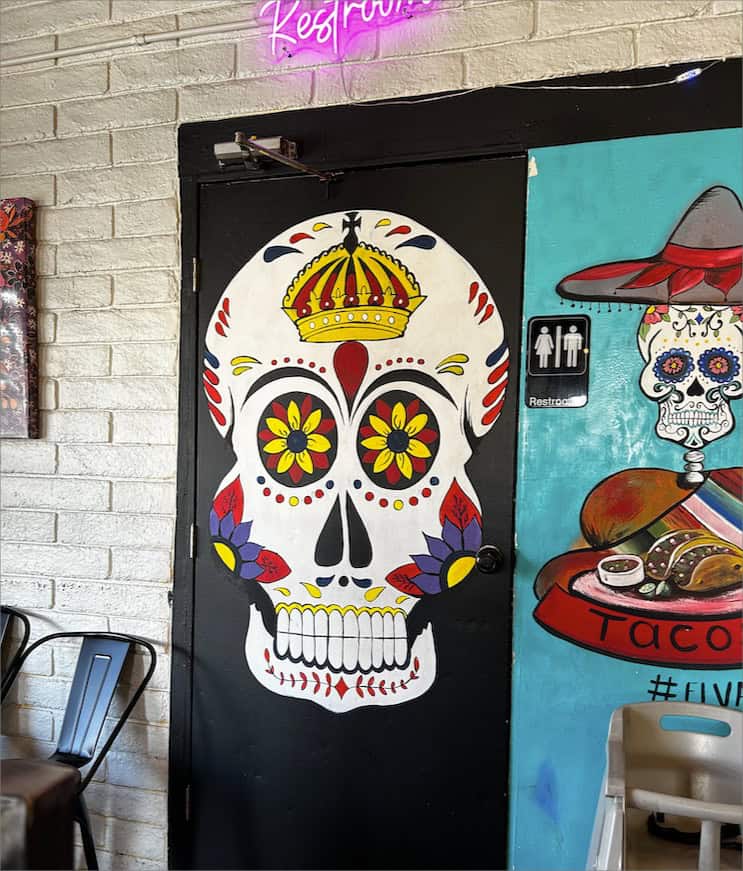 A door with a painted skull featuring a crown and colorful designs adds character to El Vagon Tacos. A neon "Restroom" sign glows above, chairs rest on the left, and a partial mural and restroom sign are visible on the right, hinting at more eclectic decor inside this delightful restaurant.