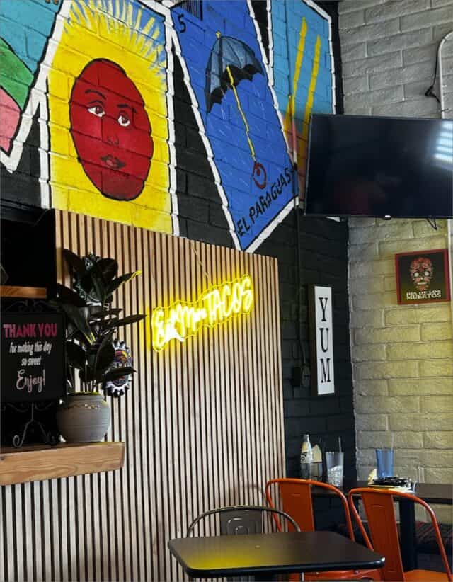 A colorful taco restaurant, El Vagon Tacos, boasts a vibrant interior with striking wall art and a neon sign reading "Eat More Tacos." There’s a TV, wooden paneling, and orange chairs around small tables adorned with remnants of delicious tacos—a perfect spot for your next restaurant review.