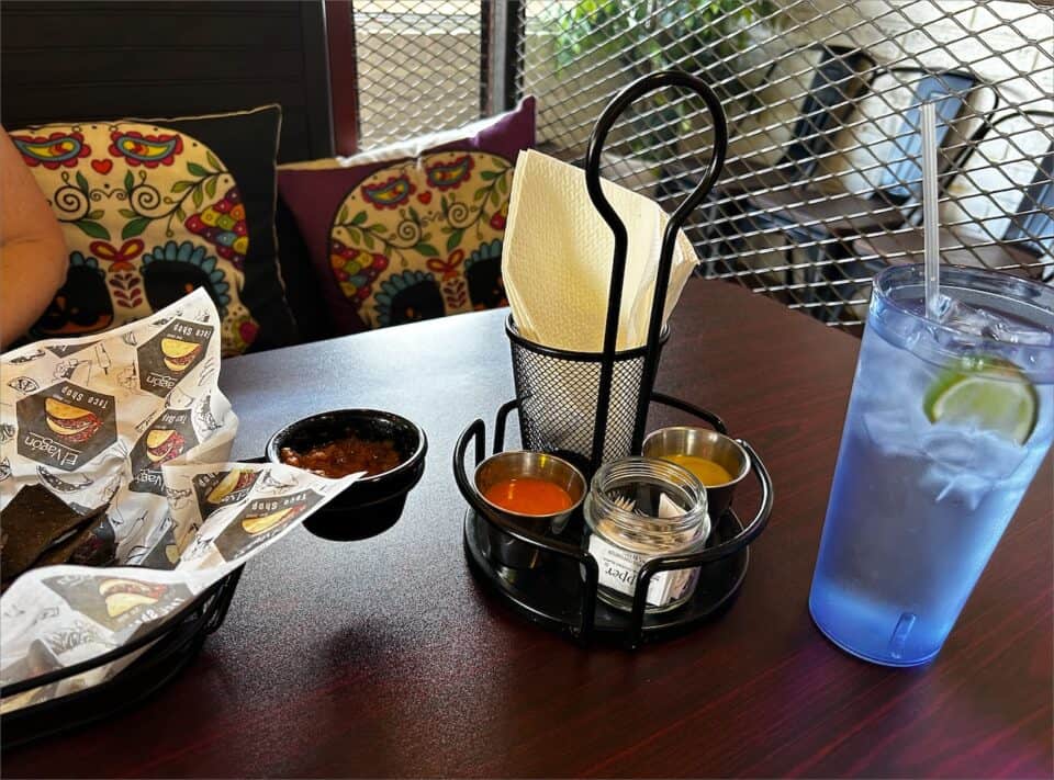 A restaurant table with a basket of chips and dip, a holder with sauces and napkins, and a blue glass of water with a slice of lime. Tacos are served on vibrant plates, complementing the colorful cushions in the background.