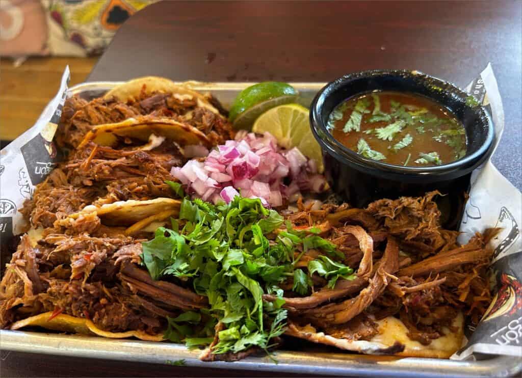 A tray of El Vagon Tacos showcases perfectly seasoned shredded meat, topped with chopped onions and cilantro, accompanied by a fresh slice of lime and a side of dipping sauce. Restaurant Review: These tacos are a flavorful delight for any taco enthusiast!