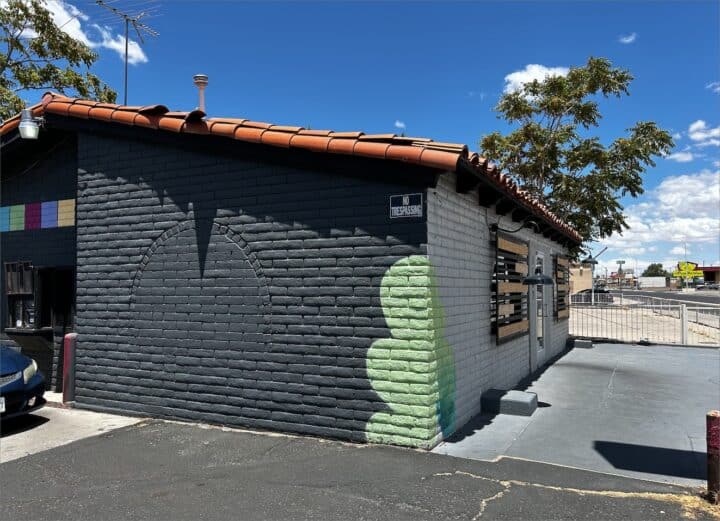 Small building with dark gray brick exterior, red-tiled roof, and a mural of a green cactus. A colorful strip adorns the upper left wall. El Vagon Tacos offers delicious fare and the surrounding area appears to be a paved parking lot—perfect for easy access when picking up your taco fix.