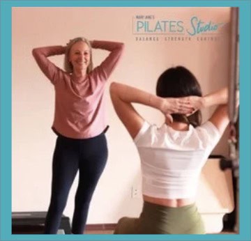 Two women, facing a wall mirror, perform an exercise with hands behind their heads at Mary Jane's Pilates studio.