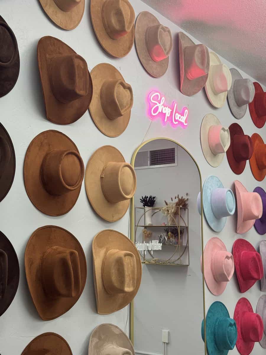 A vibrant wall display of various colored hats sits next to a mirror, while above, a neon "Shop Local" sign gleams proudly. It's as if the scene beckons you into the Ruby Hat Bar, where each shelf tells a stylish story with its unique decorations.