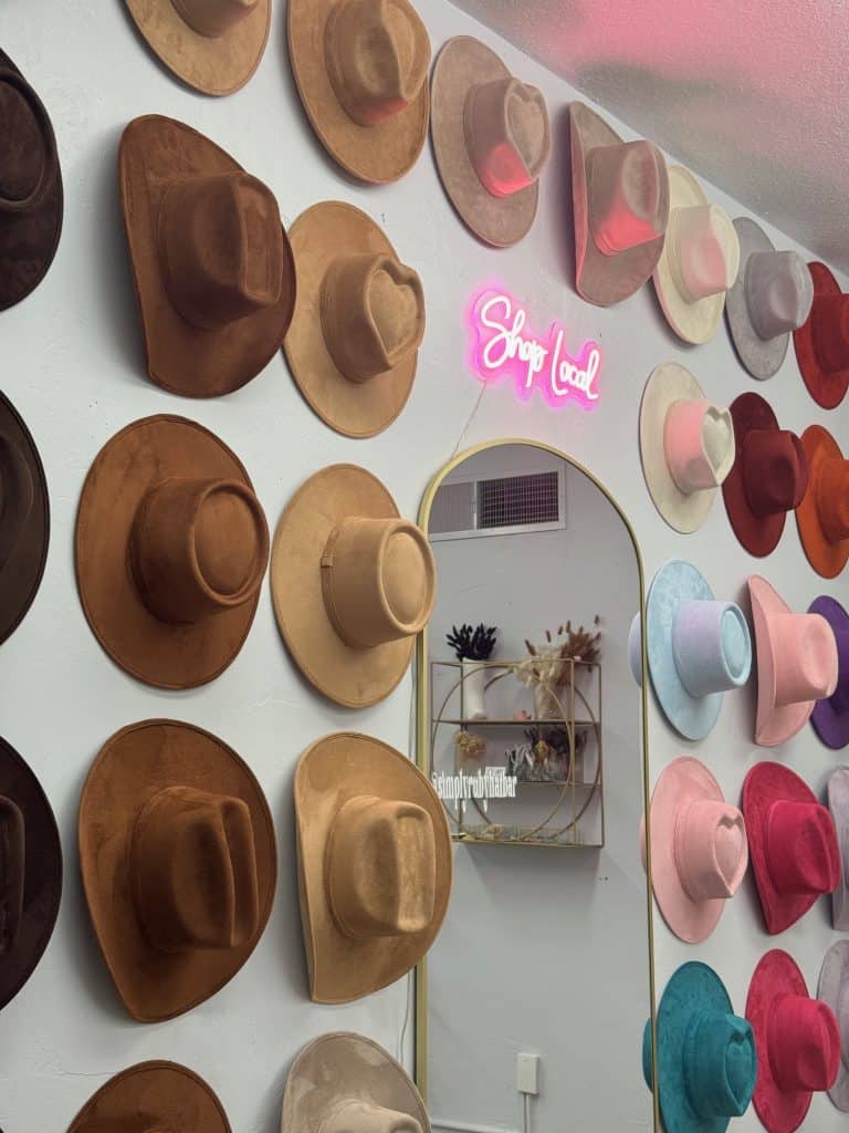 The Wall display at Simply Ruby Hat Bar features various colored wide-brimmed hats, with a neon "Shop Local" sign and a wall-mounted mirror spotlighting this charming business.