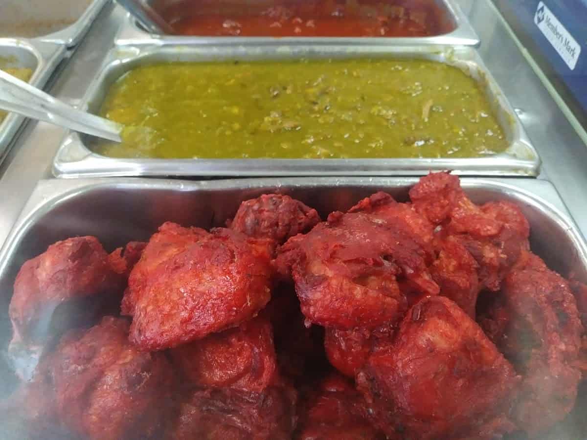 Trays of food in a buffet, featuring red fried items in the foreground and vibrant green and red curries from La Mexicana in the background, reminiscent of flavors you might find at a bustling tortilleria.