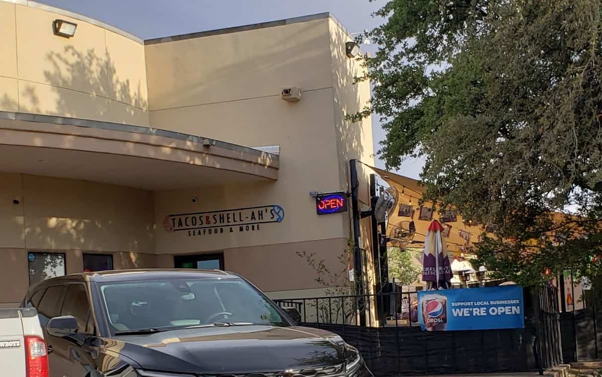 The restaurant exterior showcases the vibrant "Tacos & Shell-Ah's" sign. A neon "Open" sign glows invitingly, while a parked car is visible in the foreground beside some cozy outdoor seating.