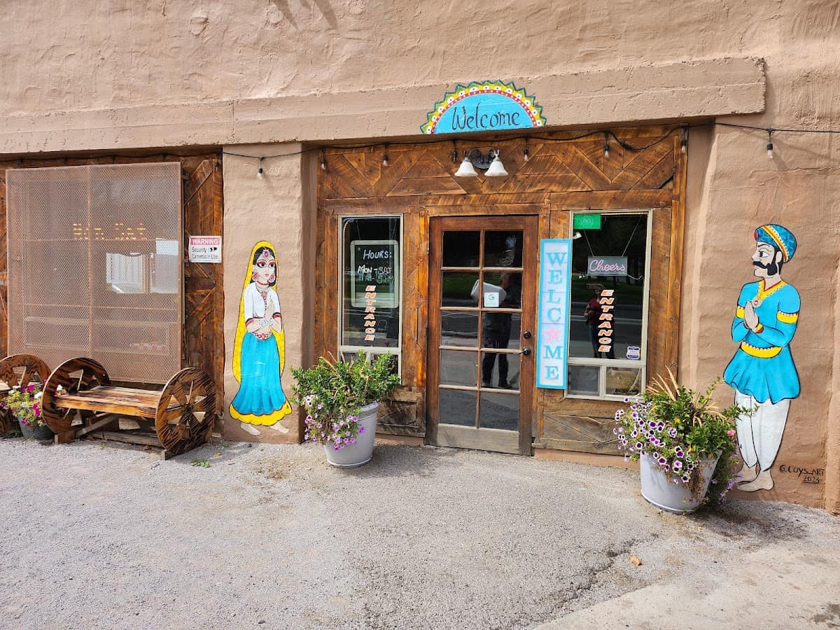 The entrance of a brown building with a wooden door greets visitors with a "Welcome" sign. Potted flowers and painted figures on either side evoke the vibrant hues of the Taste of India. A bench sits invitingly to the left, offering a space to pause and admire the scene.