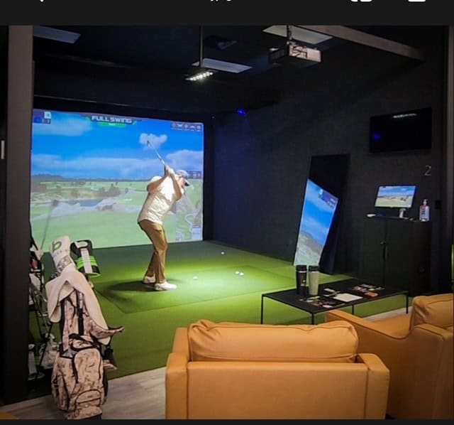 In the dimly lit room, a person immerses themselves in Back Nine Golf, skillfully swinging at an indoor simulator on vibrant green turf. Golf bags and a couch are thoughtfully placed in the foreground, creating a perfect blend of relaxation and precision.